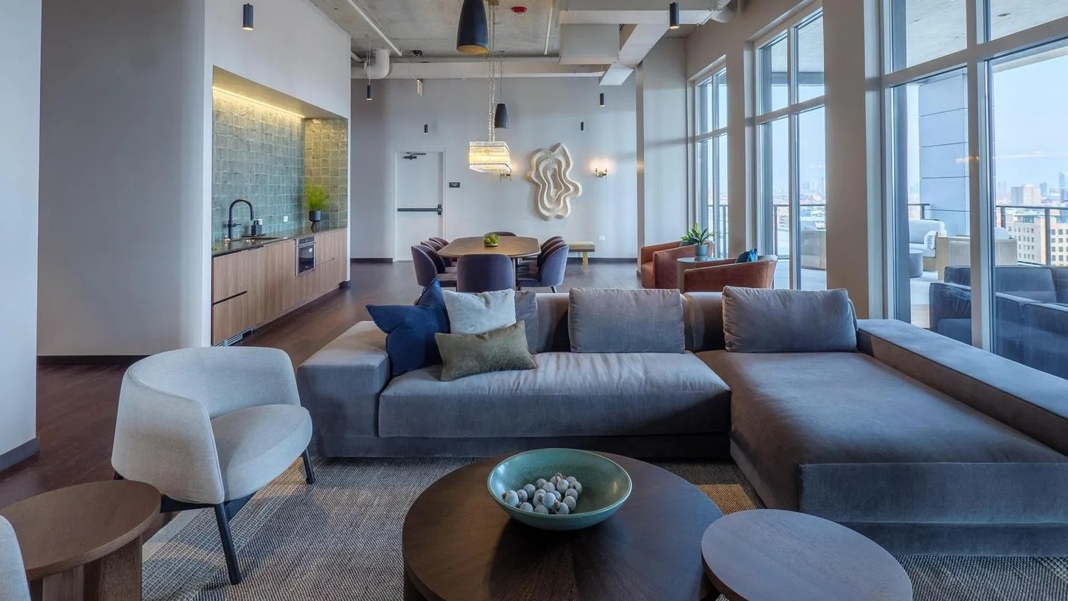 A sophisticated open-concept resident lounge with sectional sofas, armchairs, and a modern kitchen space, featuring high ceilings and city views at The Thompson at Fulton Market in Chicago