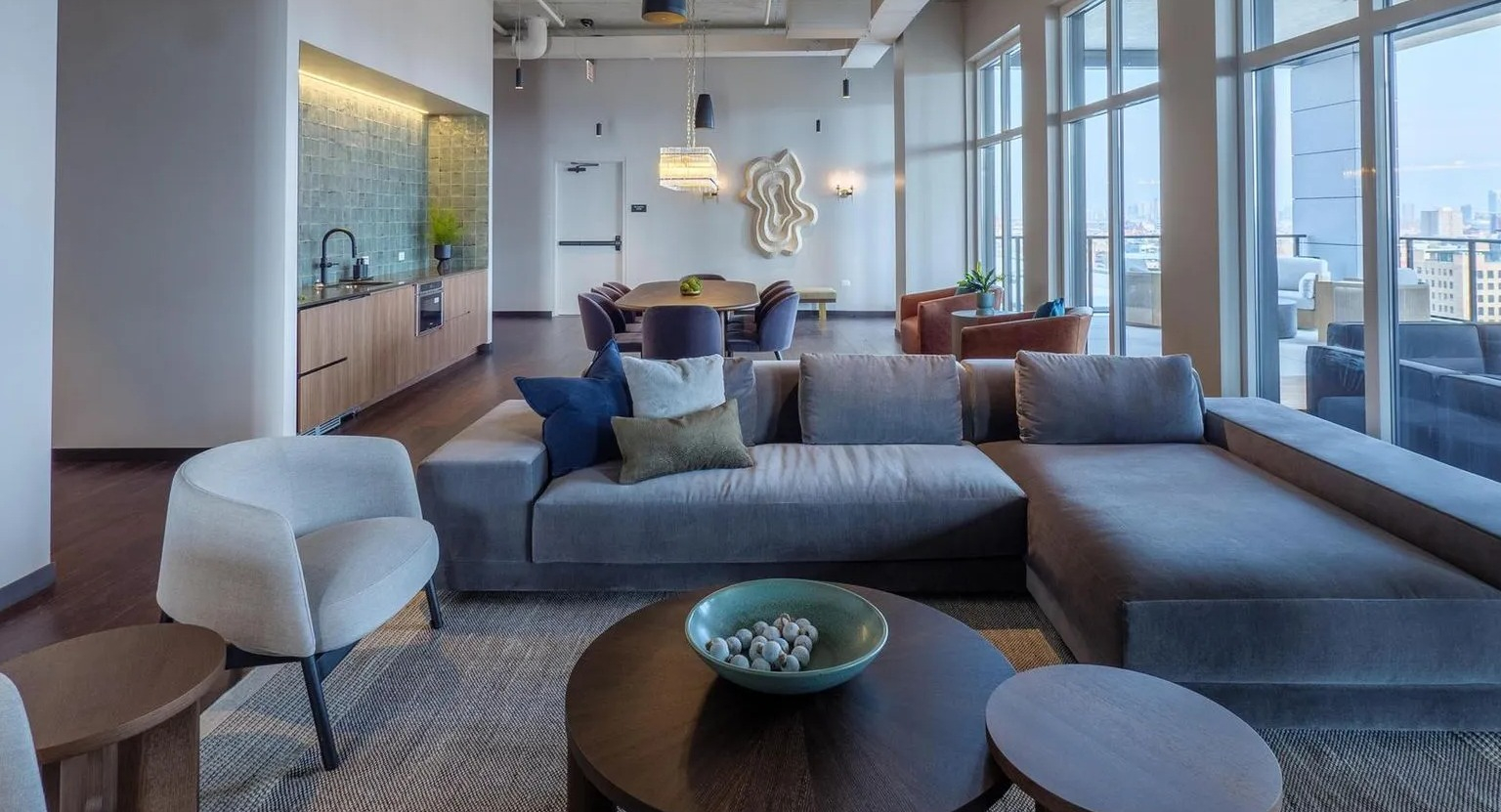 A sophisticated open-concept resident lounge with sectional sofas, armchairs, and a modern kitchen space, featuring high ceilings and city views at The Thompson at Fulton Market in Chicago