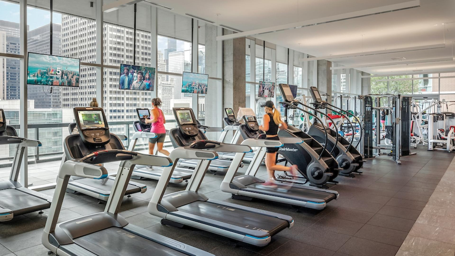 Residents exercising on treadmills and ellipticals with skyline views in the fitness center at Optima Signature apartments in Chicago