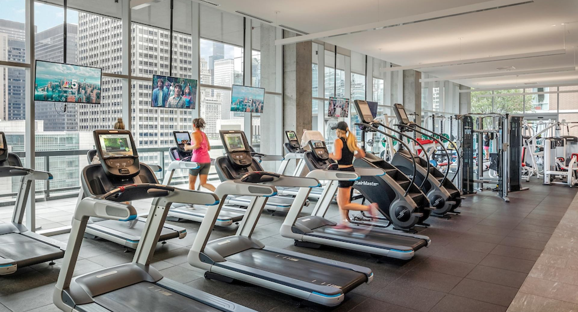 Residents exercising on treadmills and ellipticals with skyline views in the fitness center at Optima Signature apartments in Chicago