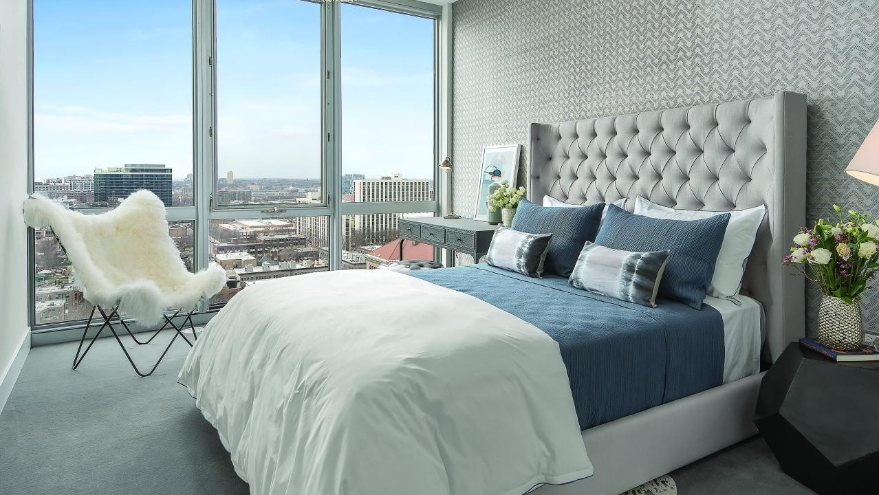 Luxurious bedroom with a tufted headboard, elegant bedding, and expansive floor-to-ceiling windows offering city views at Two West in Chicago