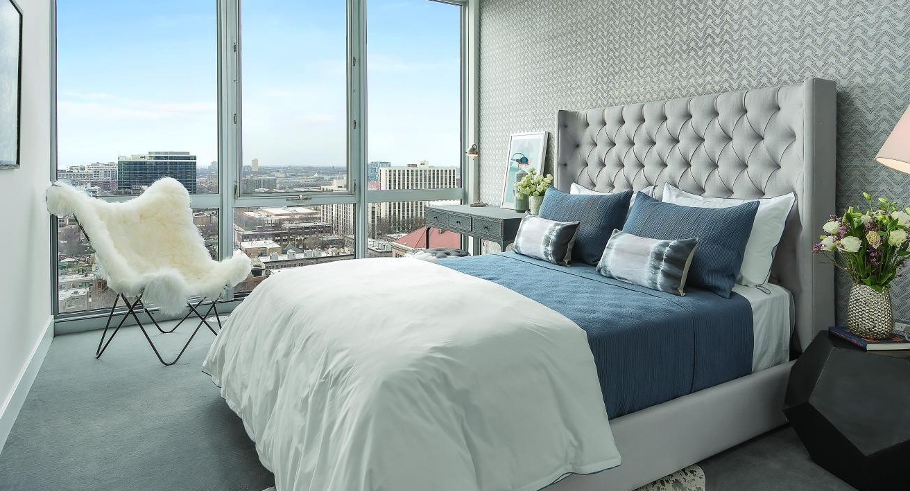 Luxurious bedroom with a tufted headboard, elegant bedding, and expansive floor-to-ceiling windows offering city views at Two West in Chicago