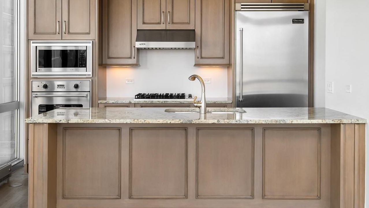 Elegant kitchen featuring custom wood cabinetry, granite countertops, a large island with an integrated sink, and stainless steel appliances at Two West apartments in Chicago