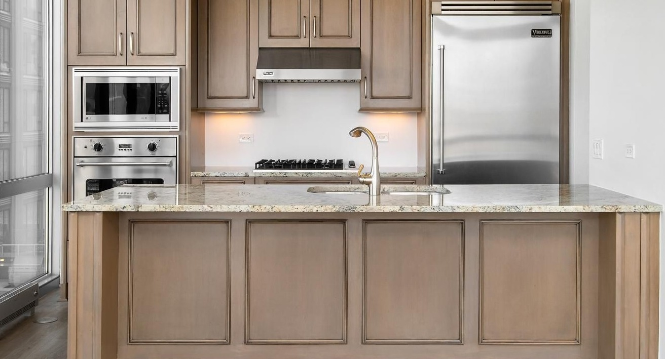 Elegant kitchen featuring custom wood cabinetry, granite countertops, a large island with an integrated sink, and stainless steel appliances at Two West apartments in Chicago