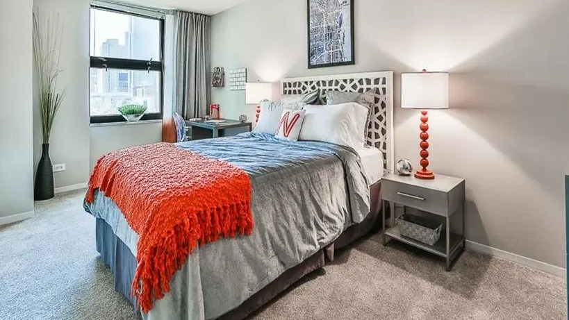Comfortable bedroom with a queen-sized bed, decorative headboard, and a desk by a window offering city views at West77 Apartments, Chicago