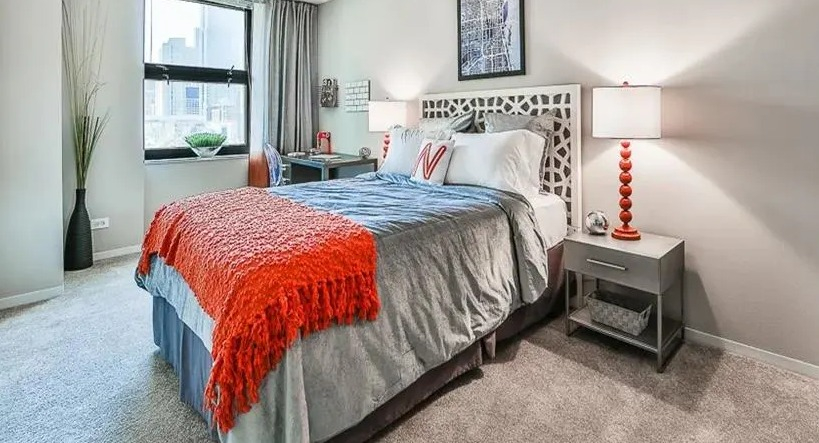 Comfortable bedroom with a queen-sized bed, decorative headboard, and a desk by a window offering city views at West77 Apartments, Chicago