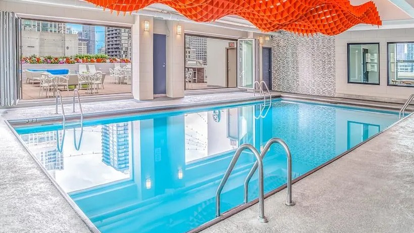 Modern indoor swimming pool with unique ceiling art and an adjacent outdoor lounge area at West77 Apartments in Chicago