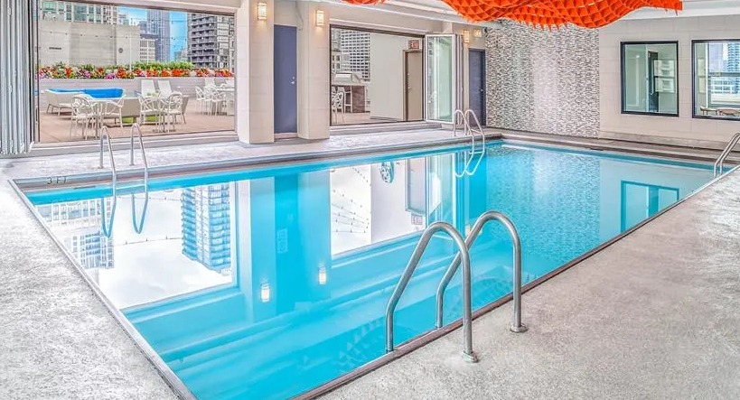 Modern indoor swimming pool with unique ceiling art and an adjacent outdoor lounge area at West77 Apartments in Chicago