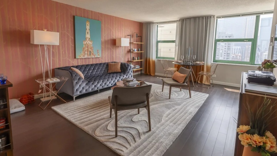 Vibrant living room with a grey tufted sofa, unique orange wall, and city views, showcasing a stylish apartment at West77, Chicago