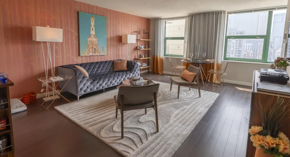 Vibrant living room with a grey tufted sofa, unique orange wall, and city views, showcasing a stylish apartment at West77, Chicago