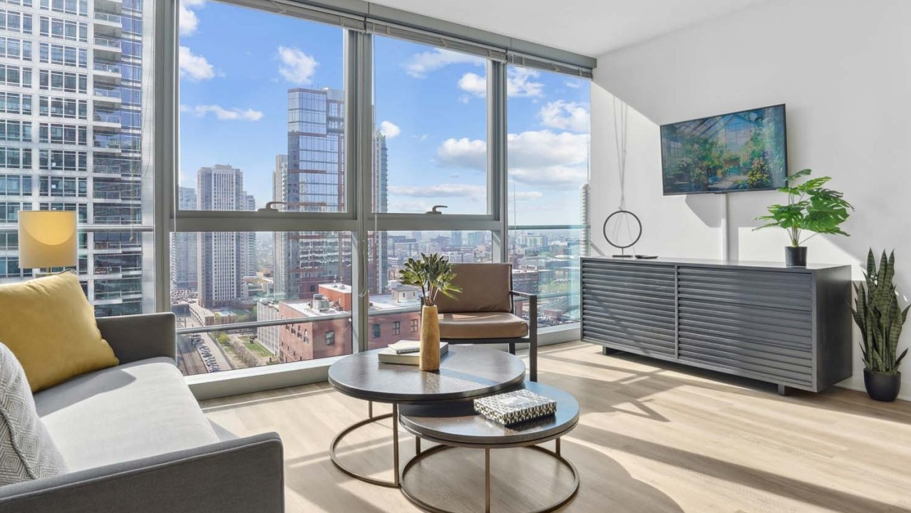 Sunlit corner living room with floor-to-ceiling windows, modern sofa and chair, nesting tables, and skyline views at Wolf Point West in Chicago