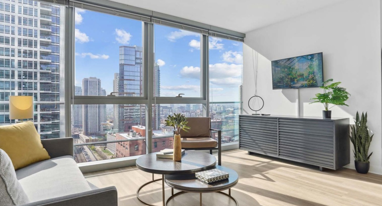 Sunlit corner living room with floor-to-ceiling windows, modern sofa and chair, nesting tables, and skyline views at Wolf Point West in Chicago