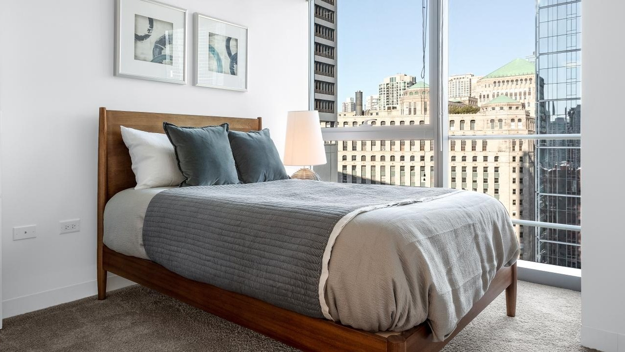 Cozy bedroom at Wolf Point West apartments in Chicago with carpet flooring, modern bed, and large city view windows