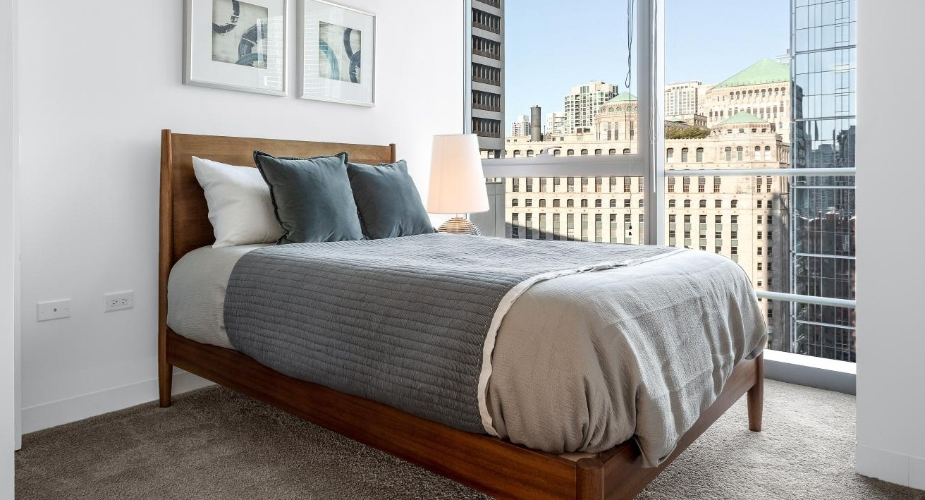Cozy bedroom at Wolf Point West apartments in Chicago with carpet flooring, modern bed, and large city view windows