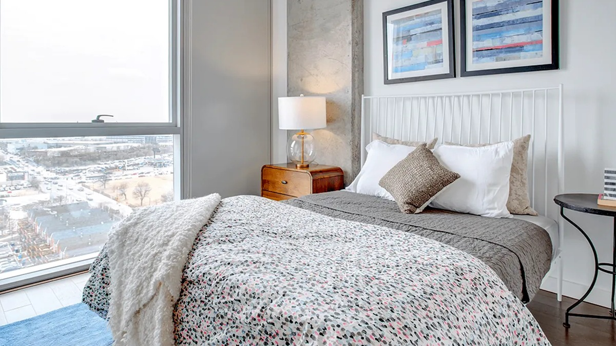 Inviting bedroom with a patterned duvet, exposed concrete ceiling, and large windows offering city views at Xavier Apartments in Chicago