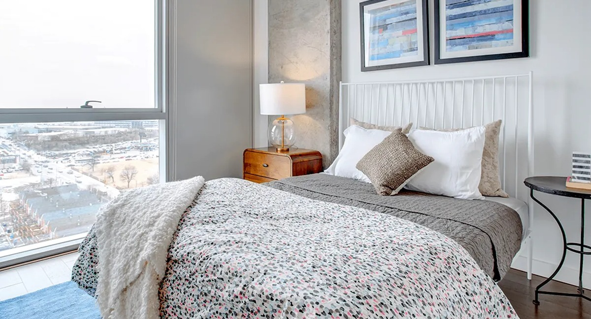 Inviting bedroom with a patterned duvet, exposed concrete ceiling, and large windows offering city views at Xavier Apartments in Chicago